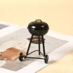 Classic black miniature kettle barbecue grill with wheels, round lid, mesh grate shelf, and tripod stand design for dollhouse garden or patio.