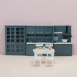 Three-piece dollhouse kitchen set in dark teal featuring a glass-door display cabinet, L-shaped counter unit with sink and cooktop, and upper cabinets with range hood, complemented by white dining furniture.