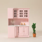 Miniature pink corner kitchen cabinet featuring white countertop, glass-paneled upper cabinets, gold faucet fixtures, and lower storage units in a charming pastel design.